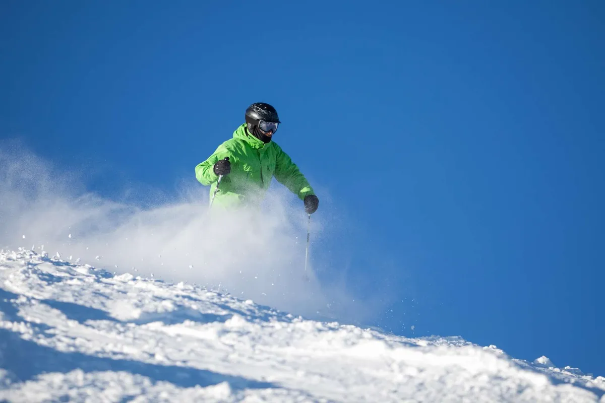 A skier in a bright green jacket carves down a snow-covered slope against a striking blue sky, with mountain views and an atmosphere brimming with winter excitement.