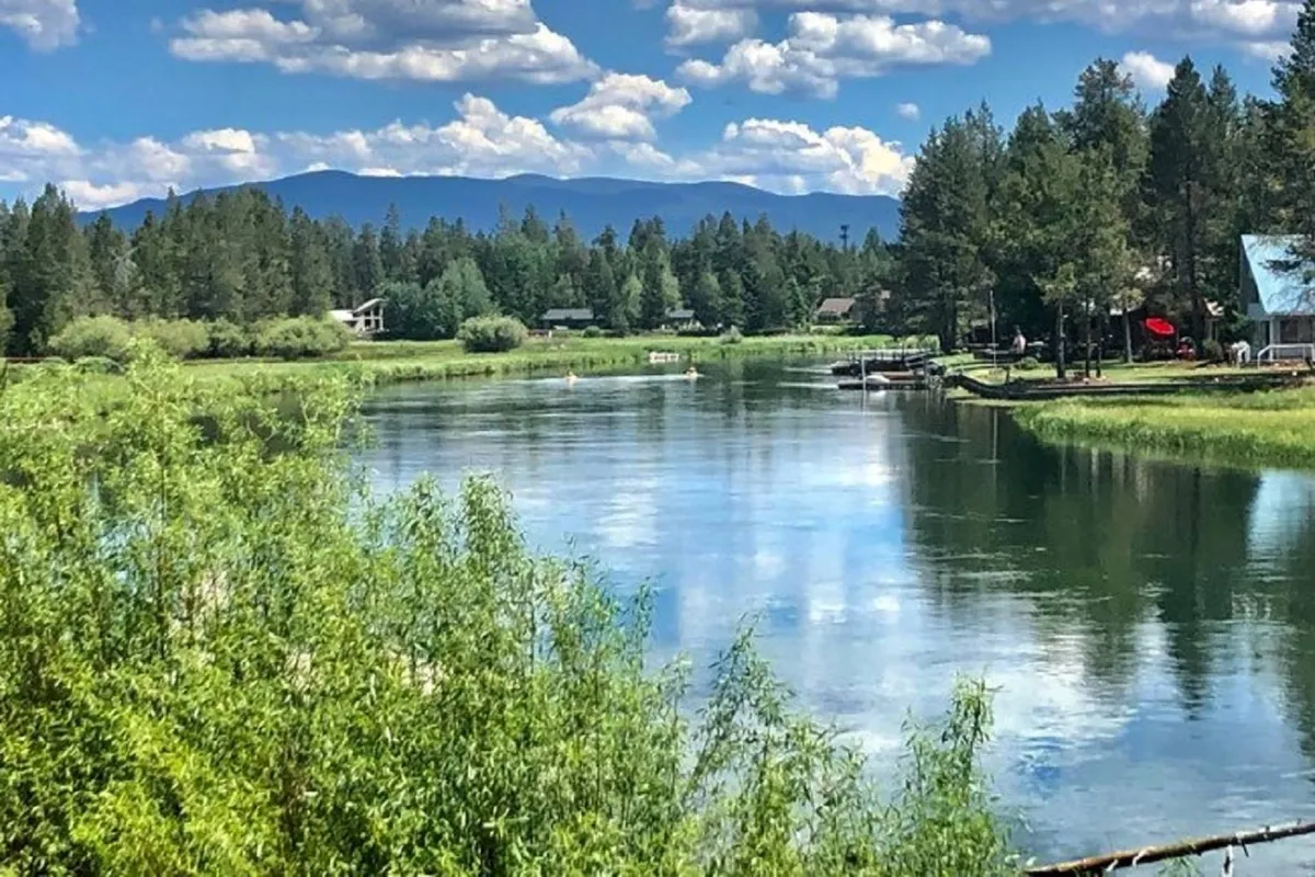 A sweeping view of the calm river bordered by lush greenery, distant mountains, and neighboring homes along the shore. The sun is low, casting gentle light across the water, setting a peaceful and scenic tone.
