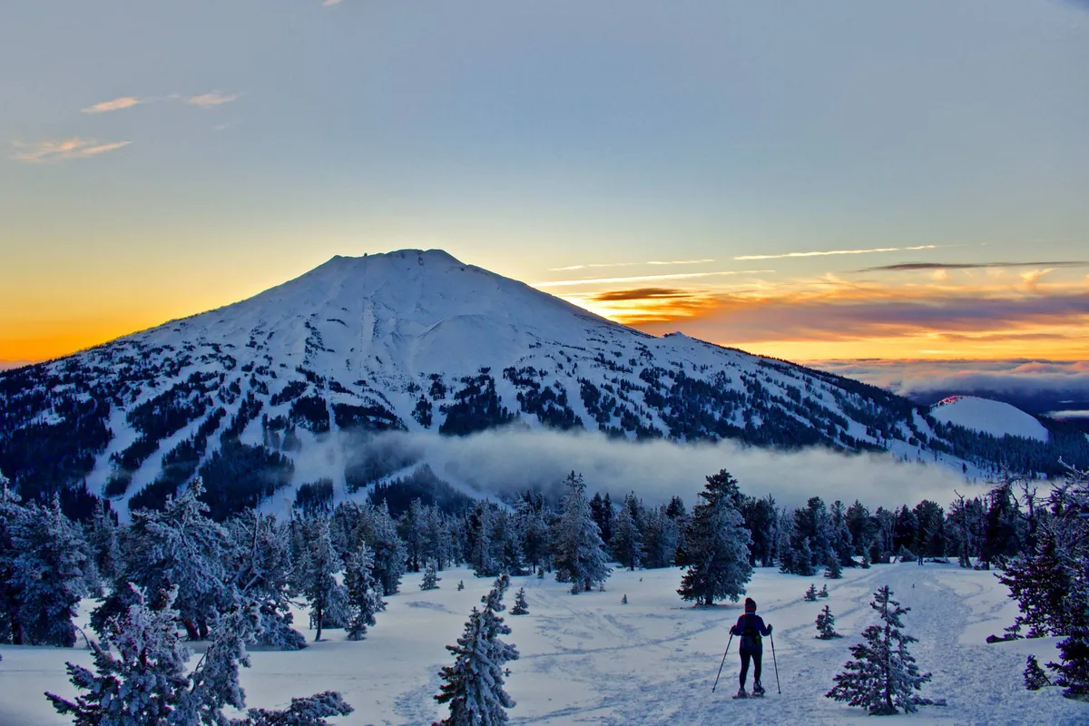 A breathtaking view of a snow-covered mountain set against a brilliant sunset sky, with snowy trees in the foreground and the silhouette of a skier carving down the slope. The image exudes both the thrill and serenity of winter sports and mountain adventures.