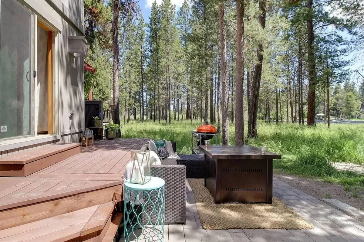 An inviting outdoor seating area on a spacious deck, featuring a grill and comfortable furniture, surrounded by a wooded backdrop. Steps connect the deck to the house, and the scene radiates a casual, serene atmosphere perfect for gatherings or quiet moments.