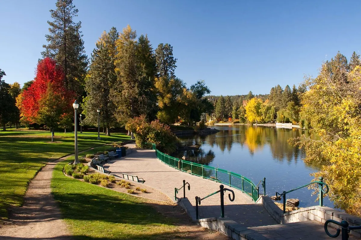 This image captures a serene lake bordered by vibrant, colorful trees in full autumn splendor. A curved pathway and green railing invite exploration, while the tranquil water reflects the foliage and sky, creating a picturesque, inviting scene. Signs of wildlife and opportunities for quiet outdoor activity are present, evoking the heart of fall experiences.
