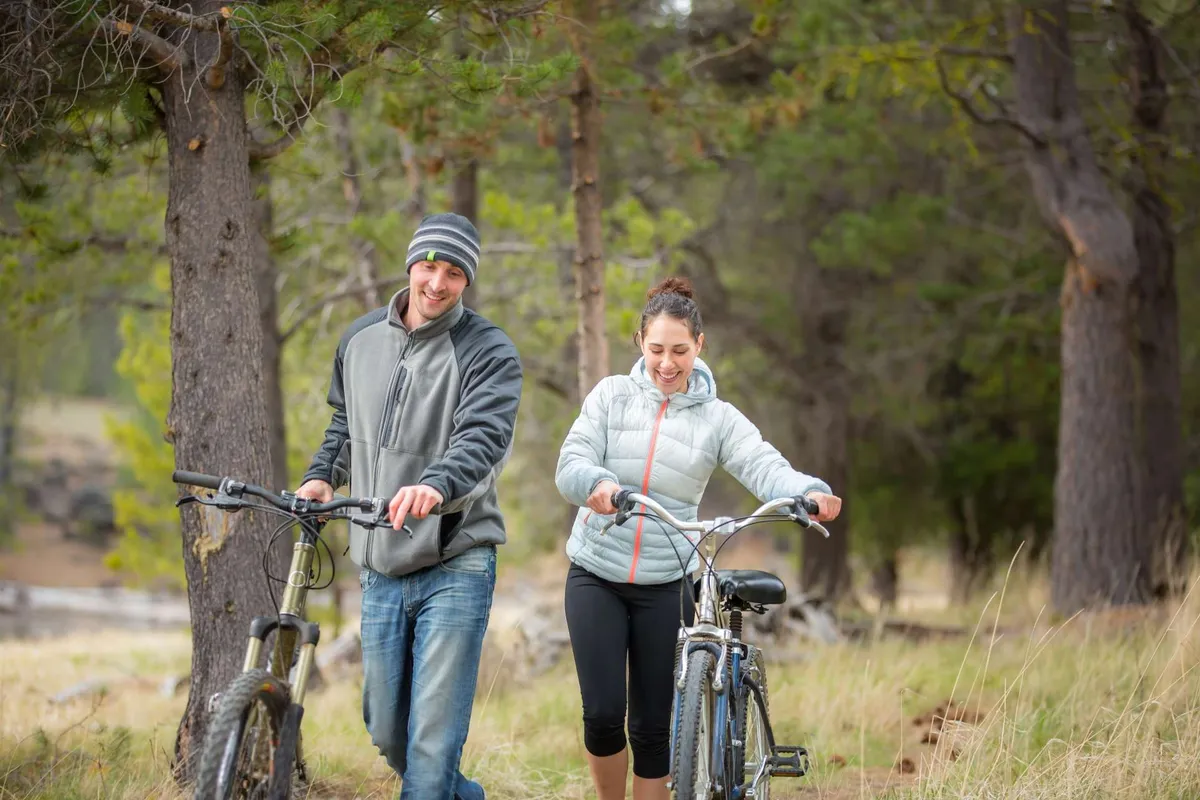 Two cyclists walk with their bikes along a scenic, wooded trail, immersed in the natural landscape. The mood is adventurous and joyful, capturing the spirit of outdoor recreation and discovery.