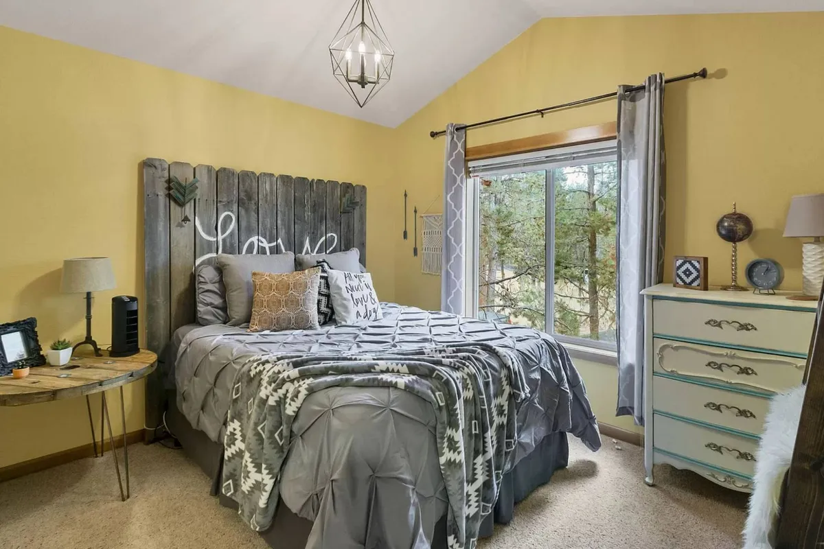 A cozy bedroom with a wooden headboard inscribed with 'Love,' decorative pillows, a large window overlooking trees, a unique light fixture, and a vintage dresser. The room bathes in soft, natural light, offering warmth and a connection to nature.