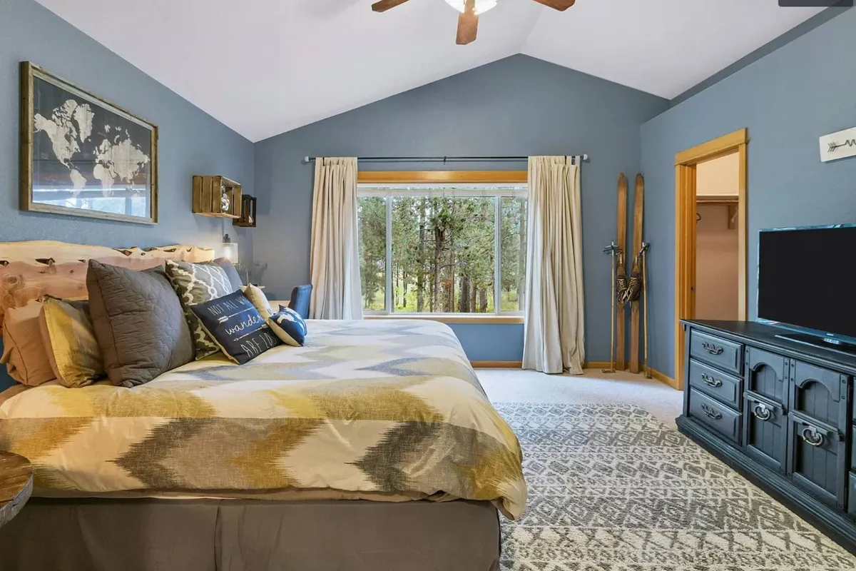 A tranquil bedroom featuring a bed with decorative pillows, a large window that floods the room with natural light, a textured rug, and warm wooden furniture. The inviting scene suggests a peaceful spot to unwind, read, or enjoy the views outside.